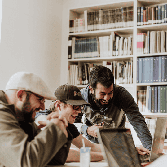 Three men collaborating and laughing while working on laptops in a library setting, emphasizing teamwork and digital marketing strategies.