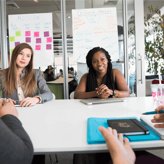 Two women engaged in a meeting, one in a blazer and the other with braided hair, discussing strategies at a table with a notebook and digital tablet, surrounded by colorful sticky notes and brainstorming materials.