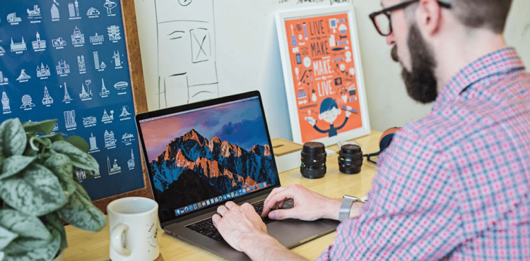 Man working on a laptop in a creative workspace, surrounded by plants, camera lenses, and inspirational posters, illustrating a dynamic digital marketing environment.