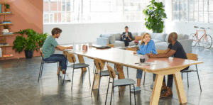 Collaborative workspace with individuals discussing digital marketing strategies at a large table, featuring plants and modern decor, reflecting a creative environment.