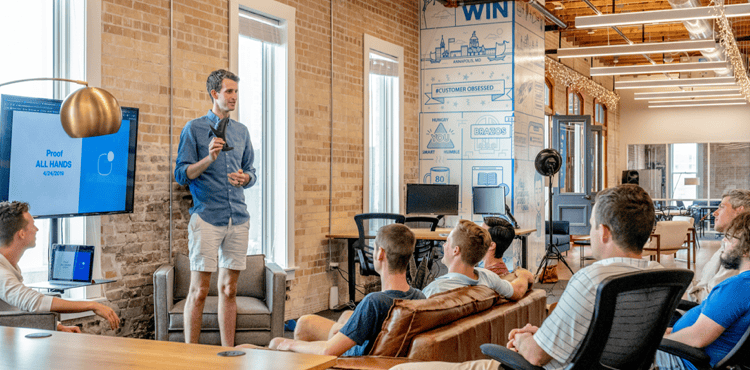Person presenting in a modern office setting, discussing insights during a team meeting, with a screen displaying "Proof ALL HANDS" and engaged audience in casual attire.