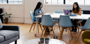 Two women collaborating at a modern workspace table, one using a laptop and the other writing, with a cozy seating area and greenery in the background, reflecting themes of design and technology in digital marketing.