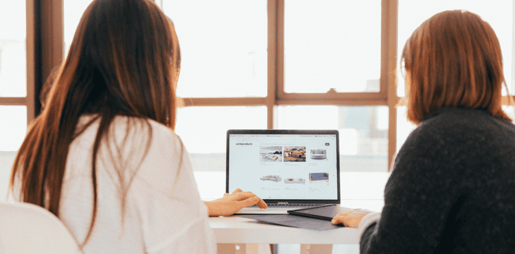 Two women collaborating over a laptop in a bright office setting, showcasing digital marketing strategies and creative design concepts.