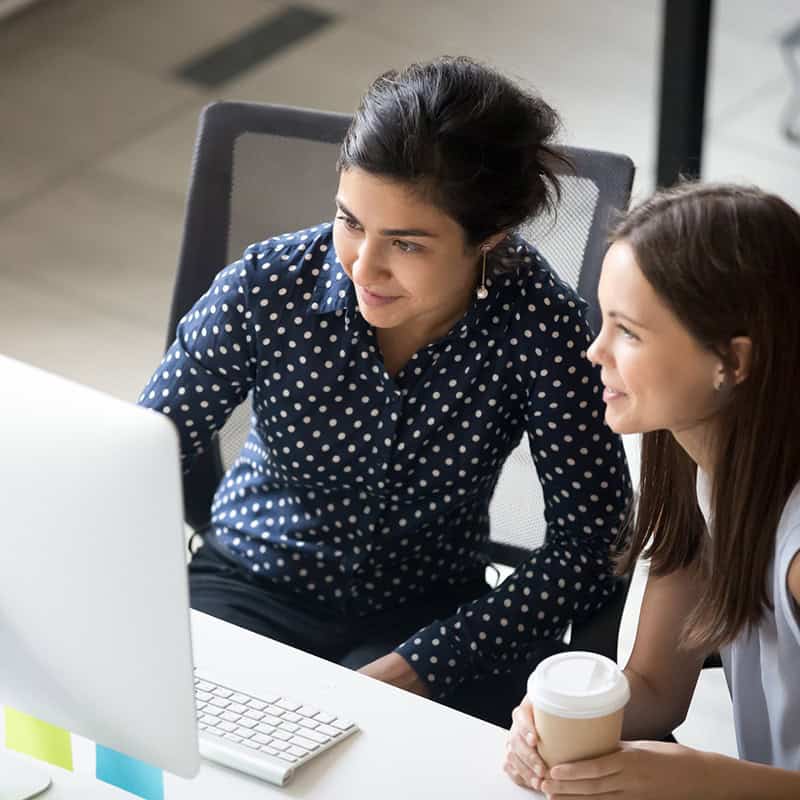 Two women collaborating at a desk, one pointing at a computer screen, discussing digital marketing strategies for local businesses and ministries.