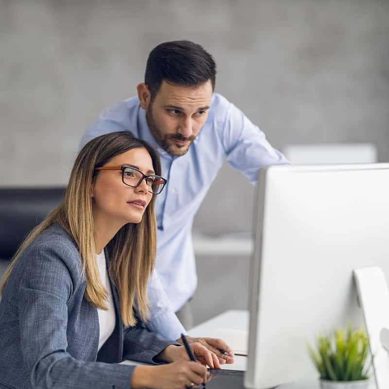 Woman writing notes while looking at a computer screen, with a man observing, in a professional office setting, emphasizing collaboration in digital marketing and website development.