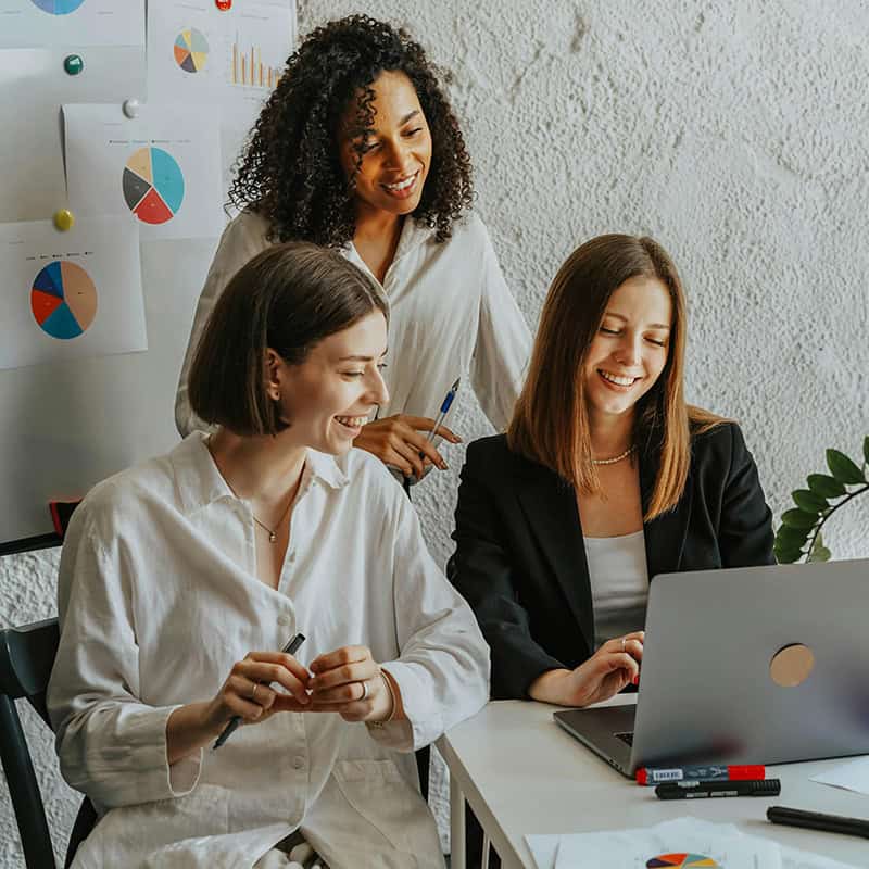Three professionals collaborating in an office setting, analyzing data on a laptop, with charts and graphs displayed on the wall, reflecting teamwork and digital marketing strategies.