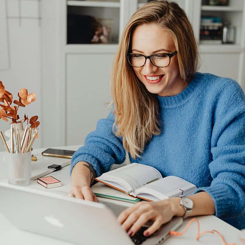 Woman smiling while typing on a laptop, surrounded by a notebook, pens, and a plant, illustrating digital marketing engagement and productivity.