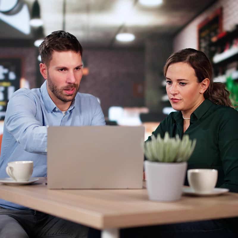 Man and woman discussing digital marketing strategies over laptop in modern café setting, with coffee cups and plant on table.