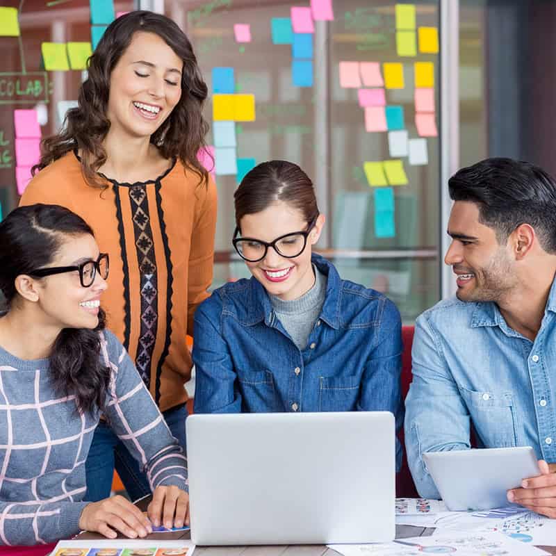 Group of four professionals collaborating during a team meeting, engaging with a laptop and discussing ideas, surrounded by colorful sticky notes on a glass wall, reflecting a creative workspace environment.