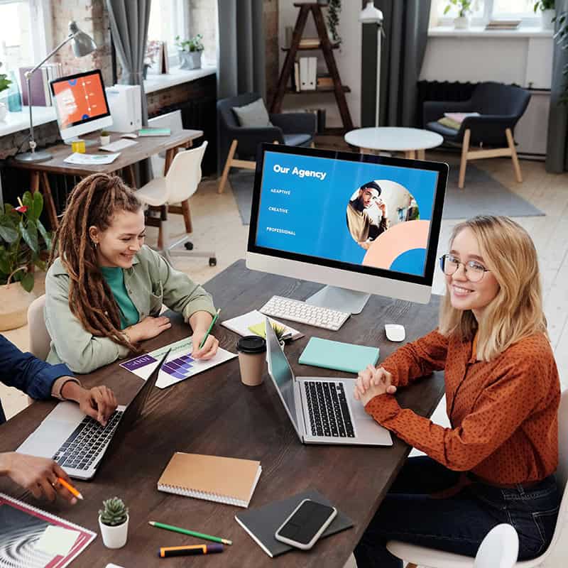 Team meeting at a creative agency, discussing strategy with laptops and colorful materials on a wooden table, featuring a computer screen displaying "Our Agency" with keywords like "Creative" and "Professional."