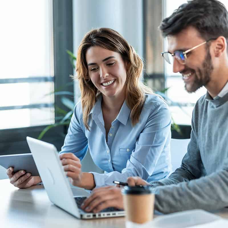 Two professionals collaborating at a desk, one using a laptop and the other holding a tablet, showcasing teamwork in a modern office environment.
