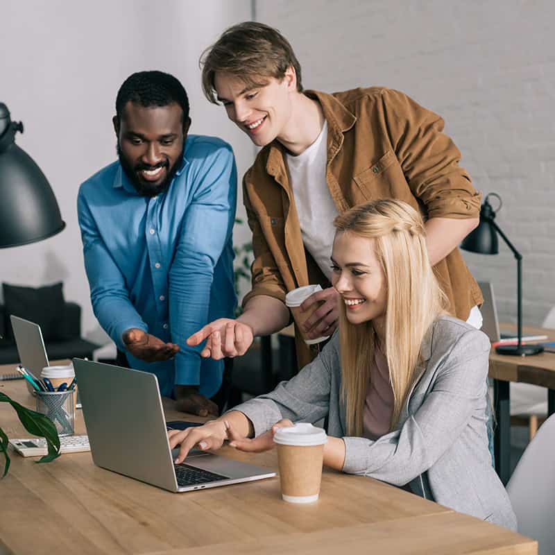 Three professionals collaborating over a laptop in a modern workspace, one woman smiling and pointing at the screen, two men engaged in discussion, showcasing teamwork and digital strategy.